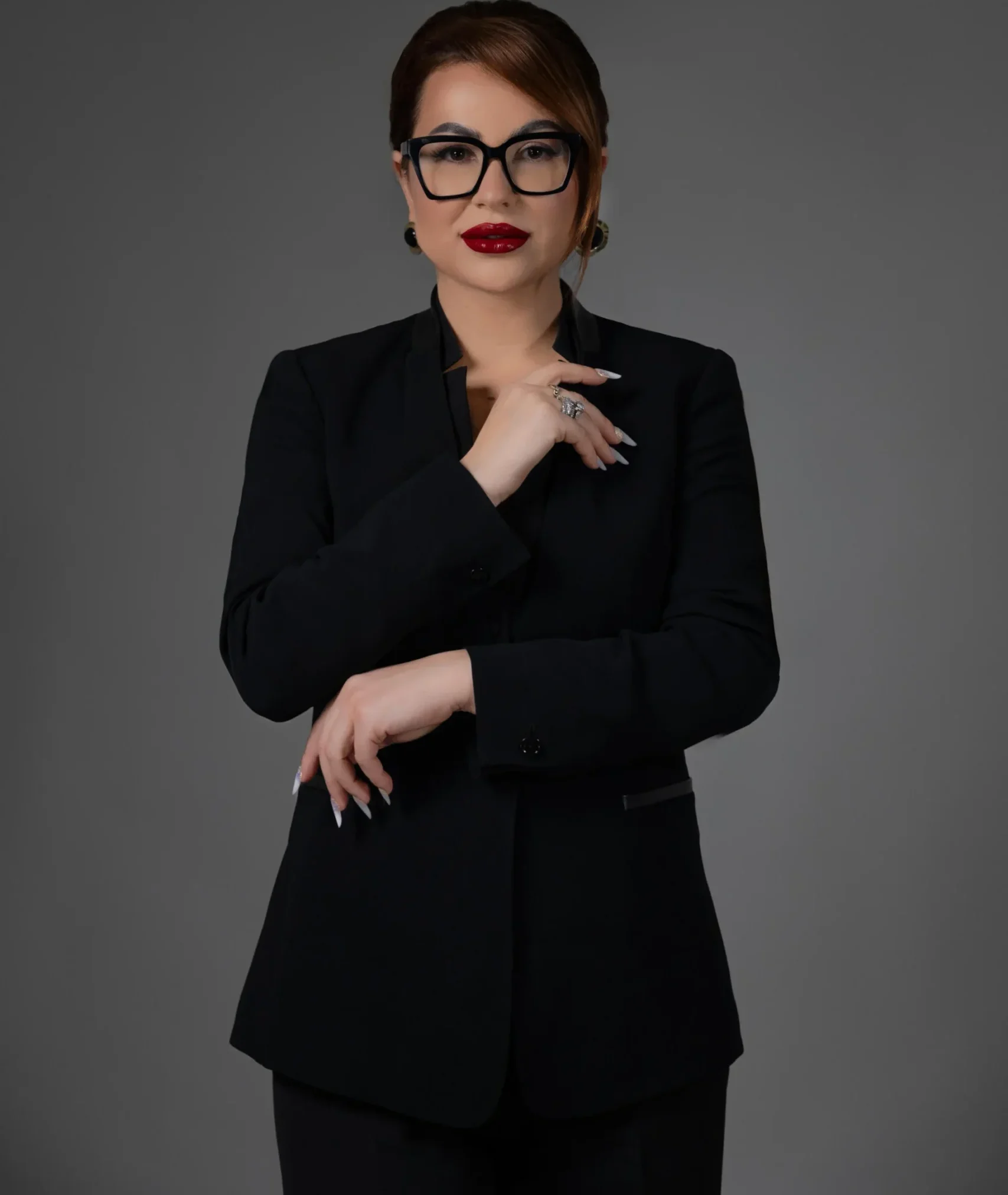 Donna in giacca nera con occhiali e rossetto rosso su sfondo grigio, posa professionale.